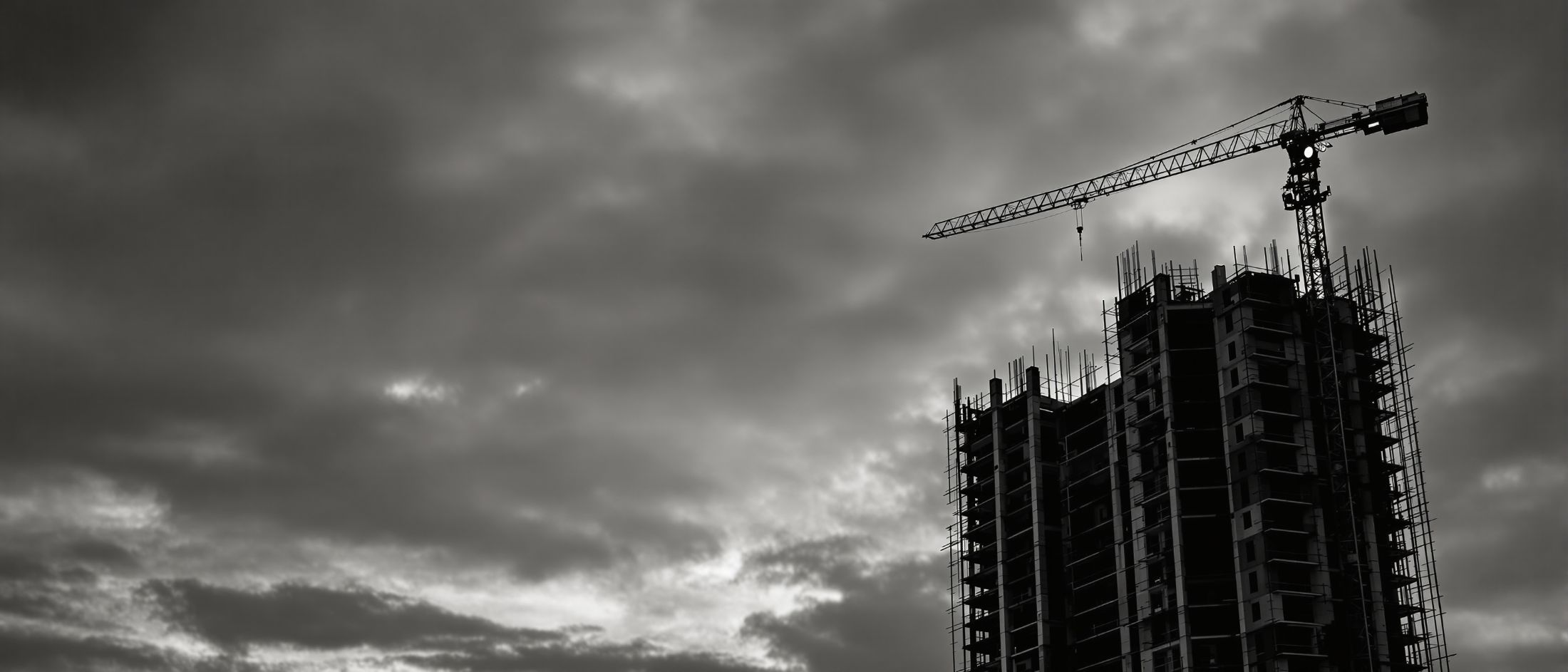 Frame of a residential building under construction at dusk, the silhouette of a
        tower crane cut against the sky.